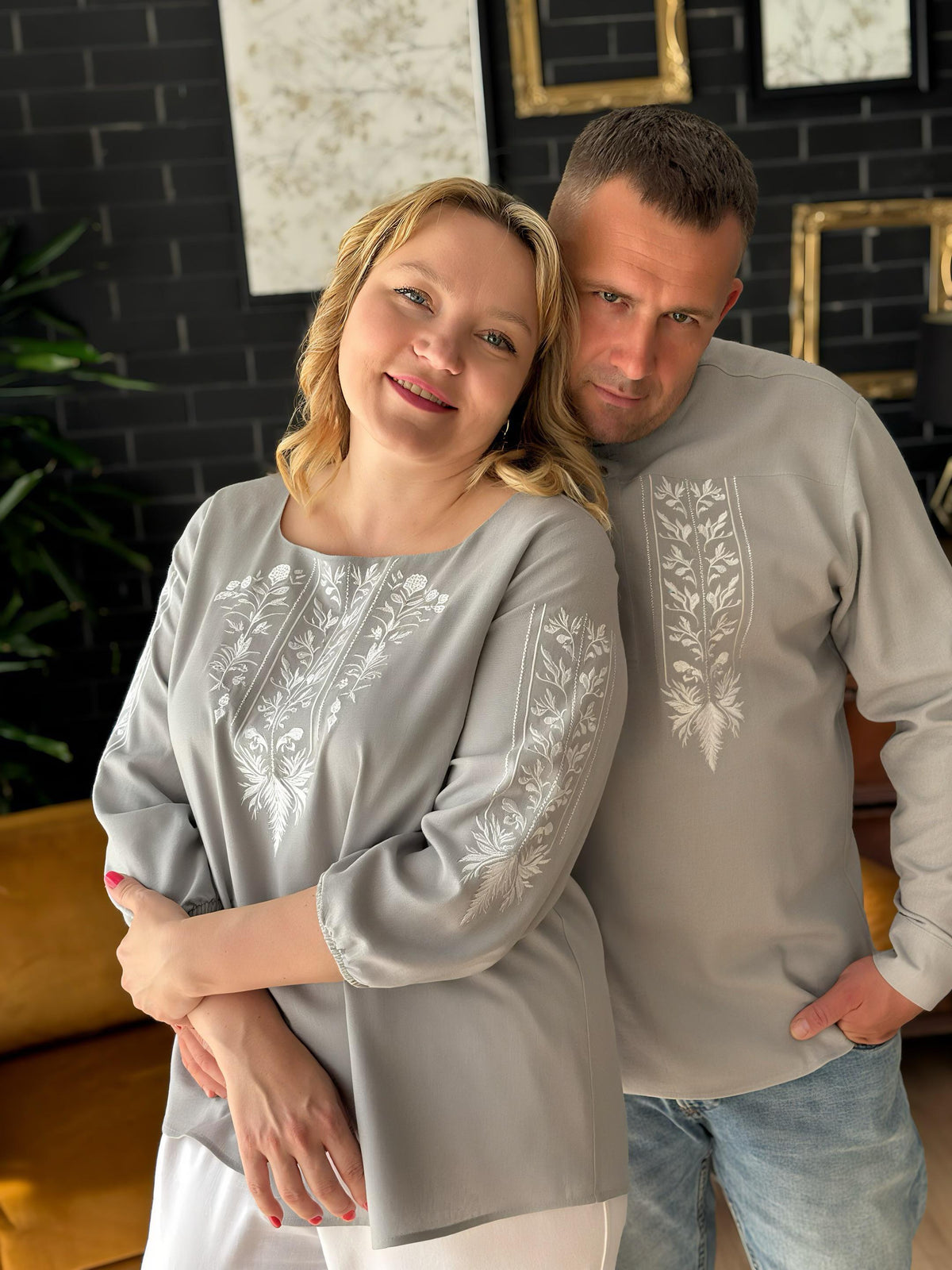 A smiling woman and a man standing close together, both wearing gray tops with white floral embroidery.